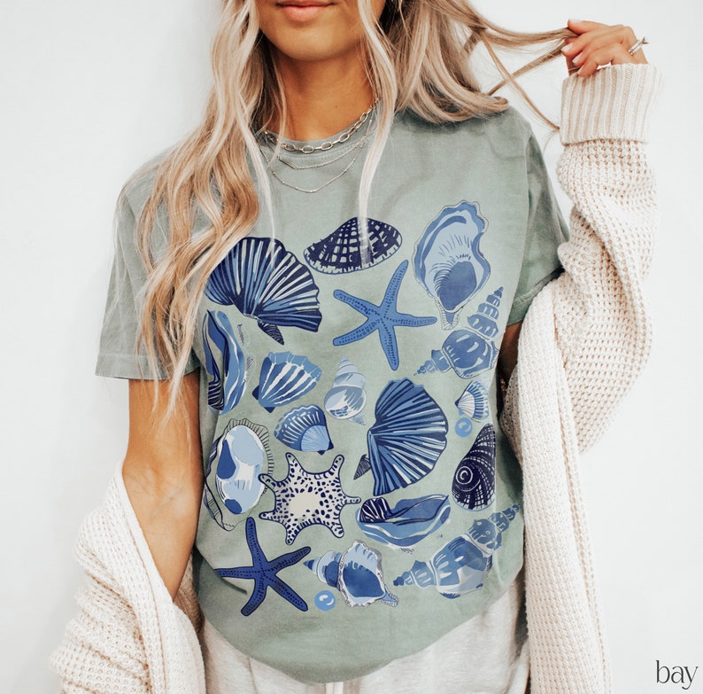Puede incluir: Una camiseta verde claro con un estampado de conchas marinas azules y blancas. La camiseta presenta una variedad de conchas, incluyendo estrellas de mar, almejas y ostras.