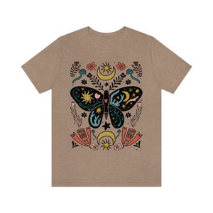 May include: A tan t-shirt featuring a black butterfly design. The butterfly has a blue and green pattern and is surrounded by stars, moons, and flowers. The design is in a folk art style.