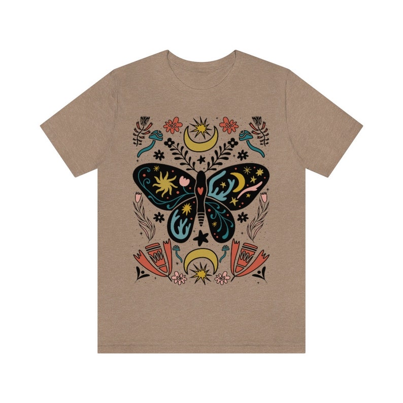 May include: A tan t-shirt featuring a black butterfly design. The butterfly has a blue and green pattern and is surrounded by stars, moons, and flowers. The design is in a folk art style.