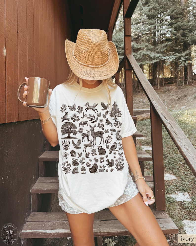 May include: Ivory-colored t-shirt featuring a detailed nature-themed graphic. The design includes various animals, plants, and trees in a monochromatic palette. The person is wearing a straw cowboy hat and holding a brown mug.