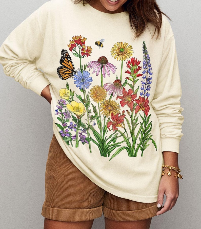 May include: A cream-colored long-sleeve sweatshirt featuring a colorful graphic print of various wildflowers, a monarch butterfly, and a small bee. The floral illustration includes red, yellow, purple, and blue blossoms with green stems and leaves.