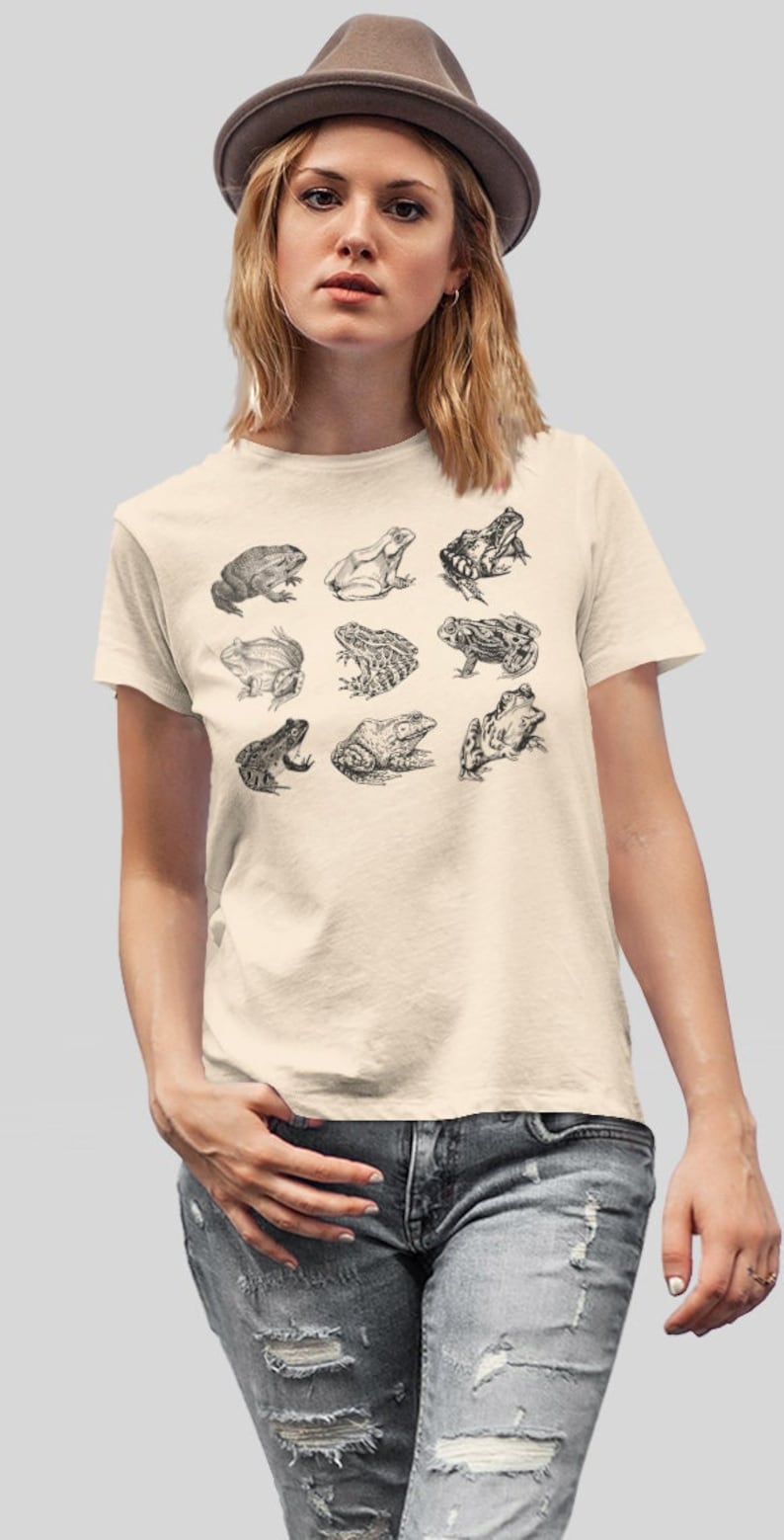 Puede incluir: Camiseta color crema con una ilustraci&oacute;n detallada en blanco y negro de nueve especies de ranas. La modelo lleva un sombrero marr&oacute;n y vaqueros desgastados. El dise&ntilde;o tiene una est&eacute;tica vintage.
