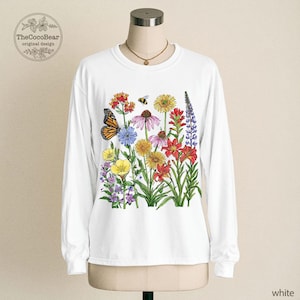 May include: A white long-sleeve crewneck shirt featuring a colorful botanical illustration of various wildflowers, a monarch butterfly, and a small bee. The graphic is centered on the chest and includes vibrant red, yellow, purple, and blue floral details.
