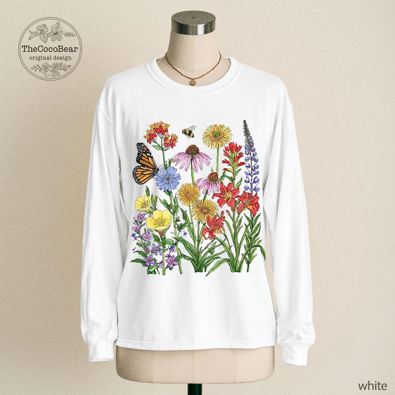 May include: A white long-sleeve crewneck shirt featuring a colorful botanical illustration of various wildflowers, a monarch butterfly, and a small bee. The graphic is centered on the chest and includes vibrant red, yellow, purple, and blue floral details.