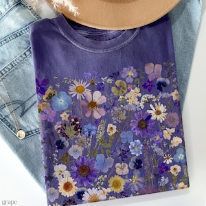 May include: A purple t-shirt with a floral print of pressed flowers in shades of purple, blue, and white.