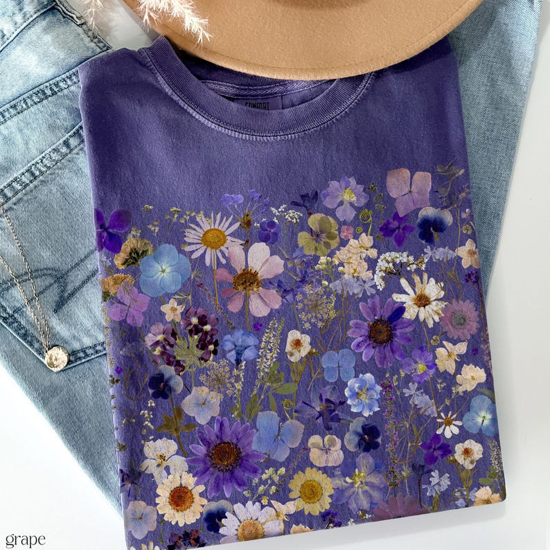 May include: A purple t-shirt with a floral print of pressed flowers in shades of purple, blue, and white.