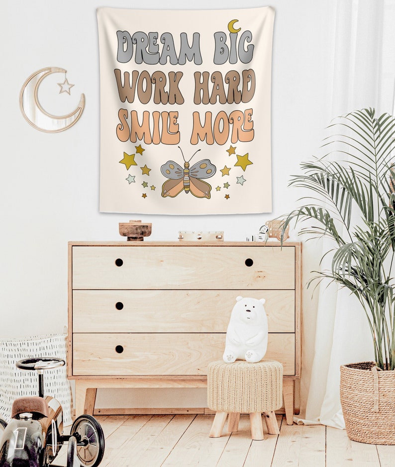 May include: A beige tapestry with the text "Dream Big Work Hard Smile More" in a retro font. A butterfly with orange, gray, and white wings is centered below the text. The tapestry is decorated with gold stars.
