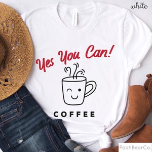 Może przedstawiać: Biała koszulka z czerwonym napisem "Yes You Can!" nad grafiką mrugającej filiżanki kawy. Słowo "COFFEE" jest wydrukowane poniżej w kolorze czarnym. Koszulka jest prezentowana ze słomkowym kapeluszem, niebieskimi dżinsami i brązowymi butami.