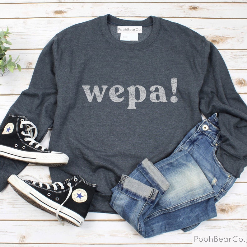 May include: A dark gray sweatshirt with the word "wepa!" printed in white on the front. The sweatshirt is laid out on a white wooden surface with a pair of black high-top sneakers and blue jeans.