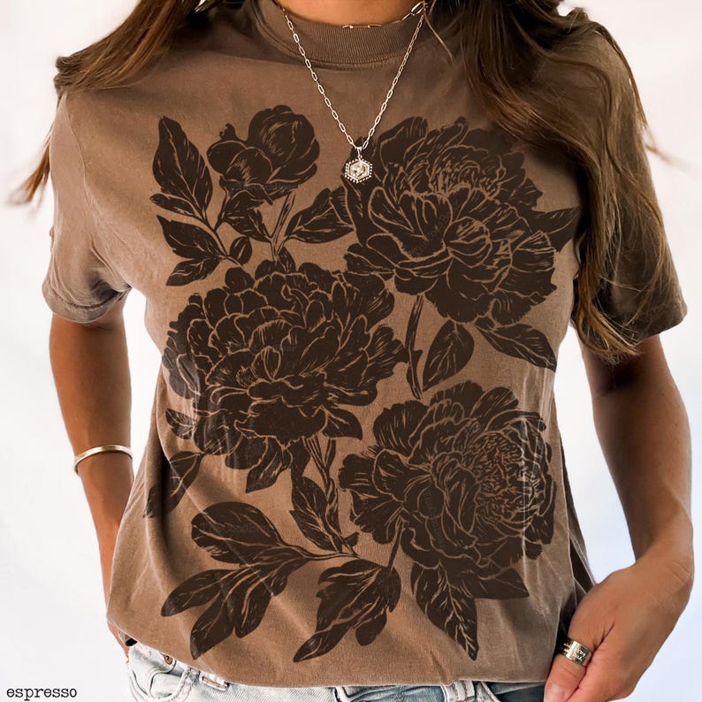 May include: Espresso-colored t-shirt featuring a large floral design in a darker brown hue. The graphic depicts detailed peony blooms and leaves. The shirt has a crew neck and short sleeves. The word "espresso" is printed at the bottom left.