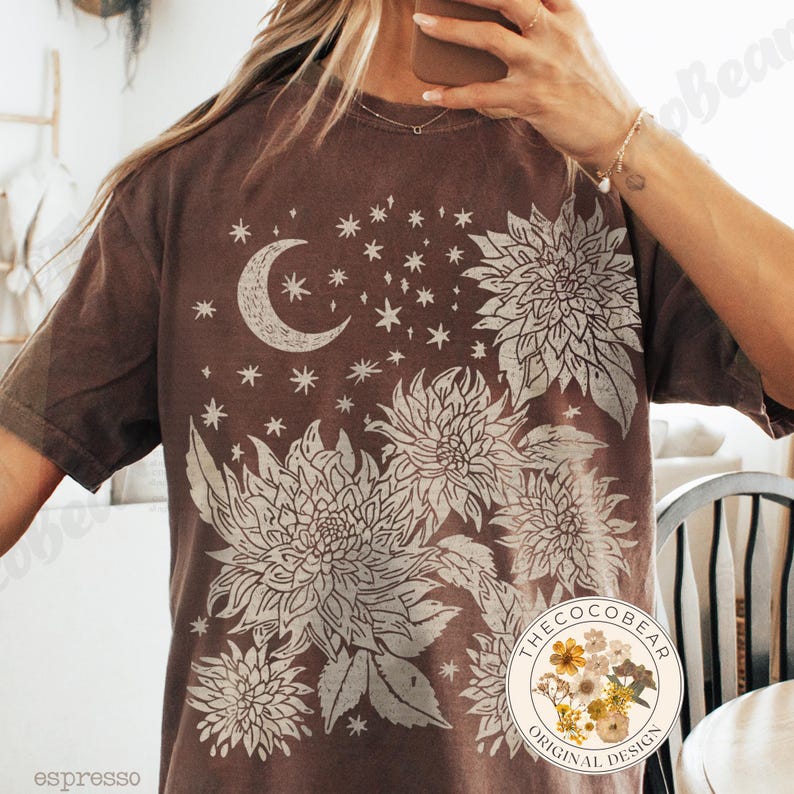 Pode incluir: Camiseta cor de caf&eacute; com um design celestial. O gr&aacute;fico inclui uma lua crescente, estrelas e grandes elementos florais em creme. A camisa tem um ajuste folgado e mangas curtas. O design &eacute; de "The Coco Bear Original Design".