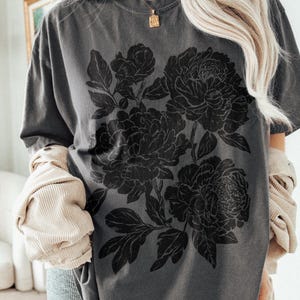 Puede incluir: Una camiseta de manga corta gris carb&oacute;n con un gran estampado gr&aacute;fico negro de tres peon&iacute;as en flor con hojas y tallos detallados. La camiseta tiene cuello redondo y un corte relajado, combinada con una chaqueta de pana beige superpuesta.