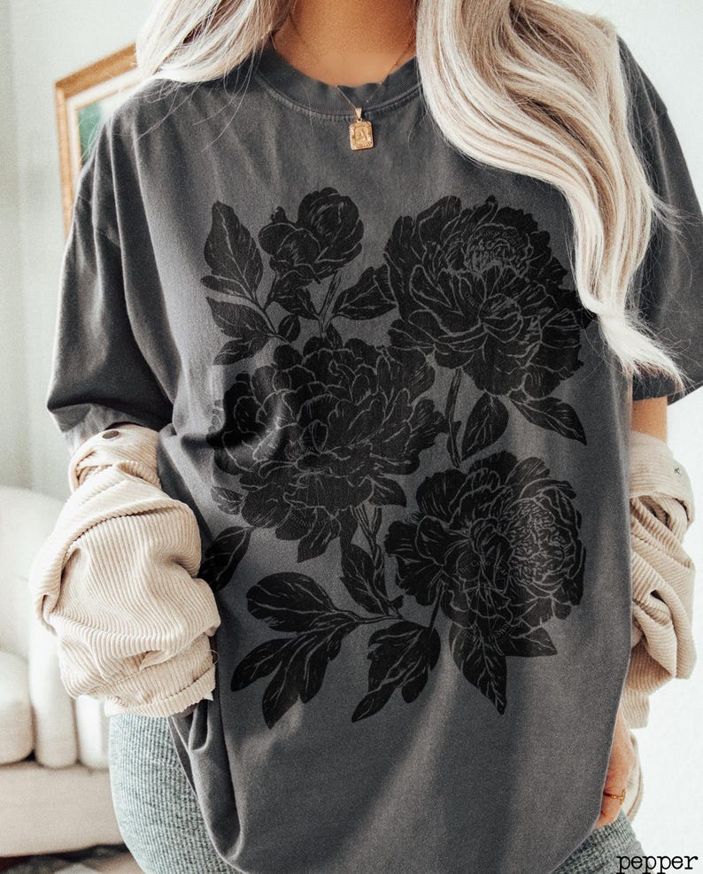 Puede incluir: Una camiseta de manga corta gris carb&oacute;n con un gran estampado gr&aacute;fico negro de tres peon&iacute;as en flor con hojas y tallos detallados. La camiseta tiene cuello redondo y un corte relajado, combinada con una chaqueta de pana beige superpuesta.