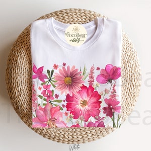 May include: White t-shirt with a colourful floral border of pink and red flowers and green leaves. The shirt is displayed on a woven, circular surface, with a tag that reads "CocoBear Design". The word "White" is at the bottom.
