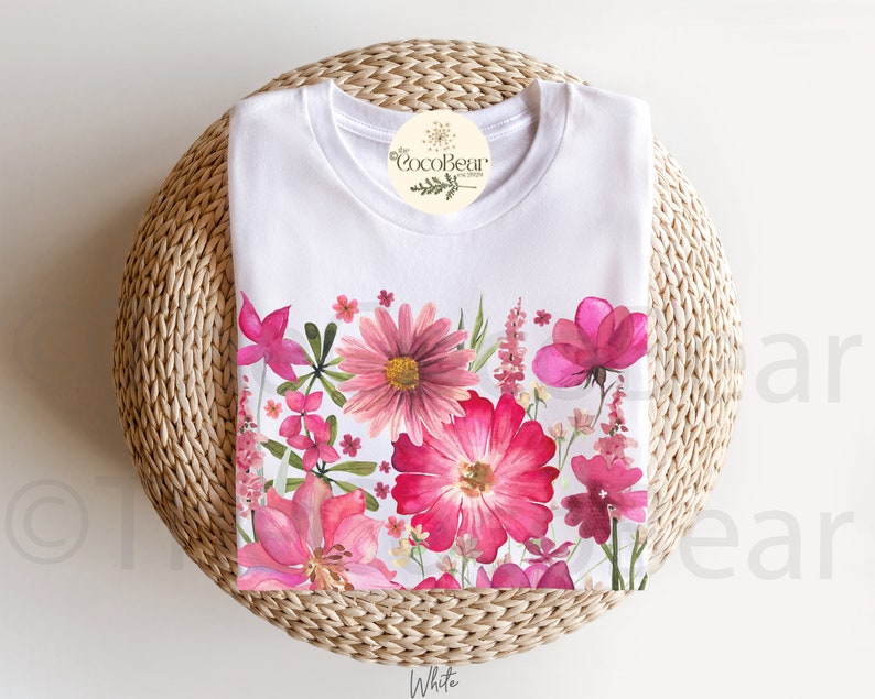May include: White t-shirt with a colourful floral border of pink and red flowers and green leaves. The shirt is displayed on a woven, circular surface, with a tag that reads "CocoBear Design". The word "White" is at the bottom.
