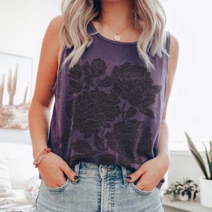 May include: A grape-colored sleeveless tank top featuring a dark, detailed floral graphic print on the front. The garment has a round neckline and is paired with light blue denim jeans. The word grape is printed in the bottom left corner.