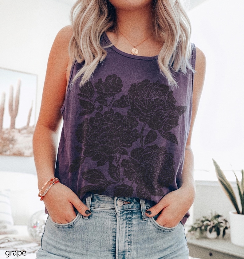 May include: A grape-colored sleeveless tank top featuring a dark, detailed floral graphic print on the front. The garment has a round neckline and is paired with light blue denim jeans. The word grape is printed in the bottom left corner.