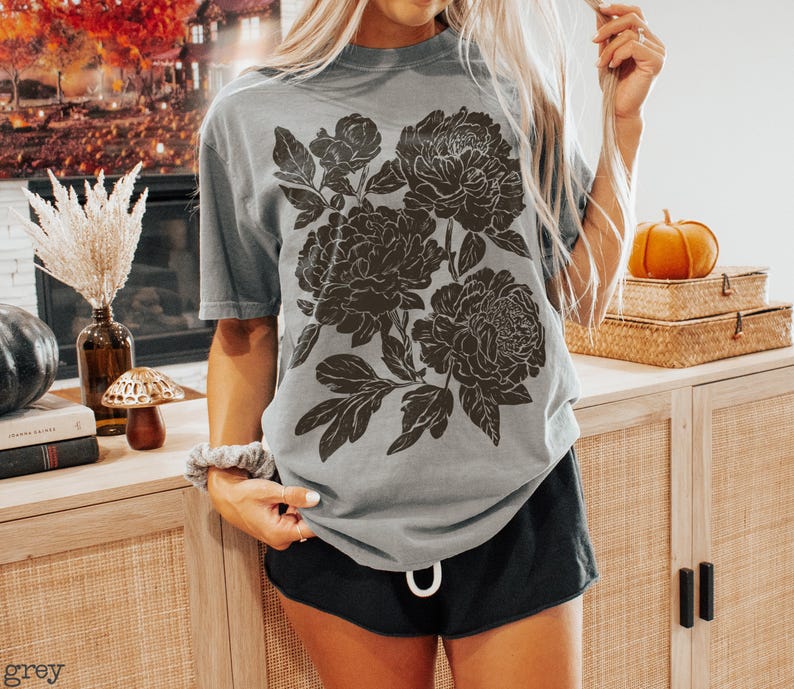 Puede incluir: Una camiseta gris de manga corta con un gran estampado gr&aacute;fico marr&oacute;n oscuro de peon&iacute;as en flor con hojas y p&eacute;talos detallados. La camiseta tiene cuello redondo y un corte relajado que muestra una ilustraci&oacute;n bot&aacute;nica en la parte frontal.