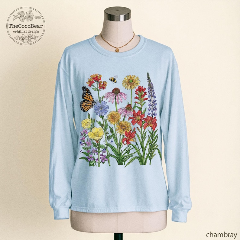 May include: A light blue long-sleeve crewneck sweatshirt featuring a colorful botanical illustration of various wildflowers, a monarch butterfly, and a small bee. The design includes red, yellow, purple, and pink flowers with green stems and leaves.