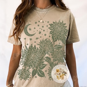 Pode incluir: Camiseta c&aacute;qui com um design floral e celestial verde. O design inclui uma lua crescente, estrelas e flores estilizadas. A camiseta tem um corte folgado e mangas curtas. Design original da The Coco Bear.