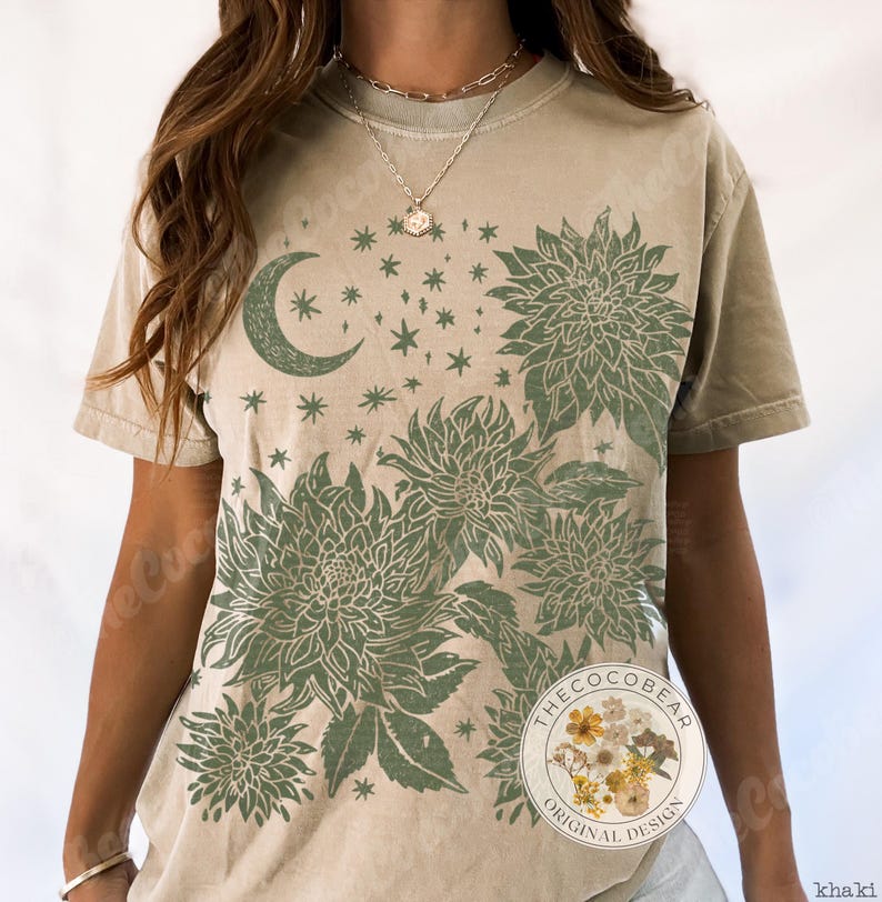 Pode incluir: Camiseta c&aacute;qui com um design floral e celestial verde. O design inclui uma lua crescente, estrelas e flores estilizadas. A camiseta tem um corte folgado e mangas curtas. Design original da The Coco Bear.