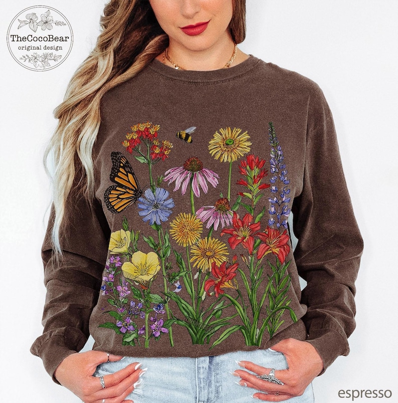 May include: A long-sleeve crewneck sweatshirt in a dark brown color featuring a colorful graphic print of various wildflowers, a monarch butterfly, and a small bee. The design includes yellow, red, purple, and orange floral illustrations.