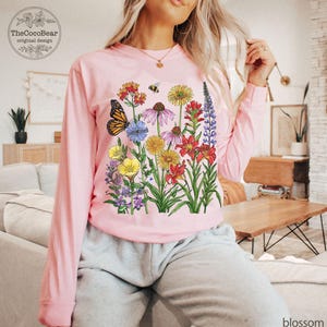 May include: A light pink long-sleeve shirt featuring a colorful graphic print of various wildflowers, a monarch butterfly, and a small bee. The design includes red, yellow, purple, and blue flowers with green stems and leaves on the front center.