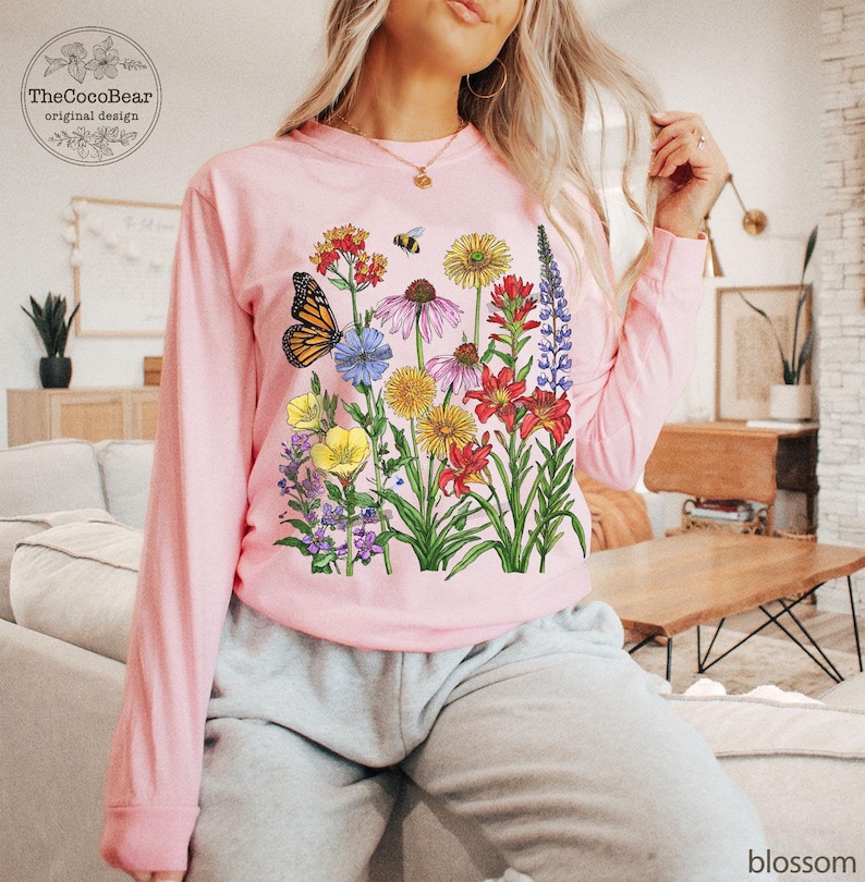 May include: A light pink long-sleeve shirt featuring a colorful graphic print of various wildflowers, a monarch butterfly, and a small bee. The design includes red, yellow, purple, and blue flowers with green stems and leaves on the front center.