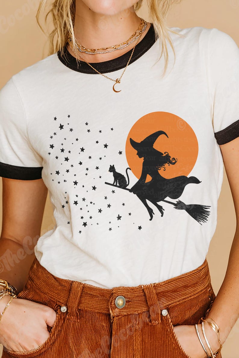 May include: Cream-coloured t-shirt with black trim, featuring a Halloween-themed design. The graphic depicts a witch on a broom with a black cat, stars, and an orange moon. A casual, everyday wear item.