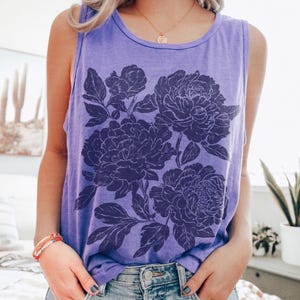 May include: A violet sleeveless tank top featuring a dark purple graphic print of large blooming peonies with detailed leaves and petals. The garment has a round neckline and a relaxed fit, with the word violet printed in the bottom right corner.