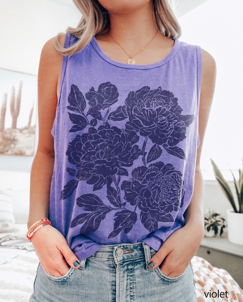 May include: A violet sleeveless tank top featuring a dark purple graphic print of large blooming peonies with detailed leaves and petals. The garment has a round neckline and a relaxed fit, with the word violet printed in the bottom right corner.