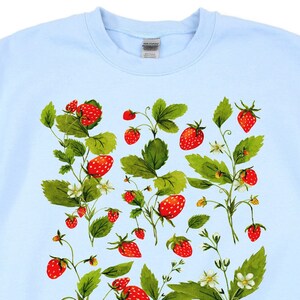 Puede incluir: Sudadera azul claro con un dise&ntilde;o de acuarela de fresas y hojas. El dise&ntilde;o incluye fresas rojas, hojas verdes y peque&ntilde;as flores blancas. Las fresas est&aacute;n dispersas por la parte delantera de la sudadera.