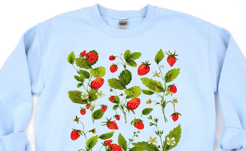 Puede incluir: Sudadera azul claro con un dise&ntilde;o de acuarela de fresas y hojas. El dise&ntilde;o incluye fresas rojas, hojas verdes y peque&ntilde;as flores blancas. Las fresas est&aacute;n dispersas por la parte delantera de la sudadera.
