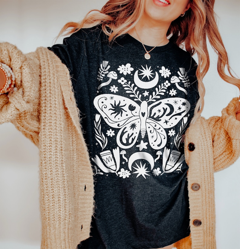 Puede incluir: Una camiseta gris oscuro con un gr&aacute;fico blanco de una mariposa rodeada de estrellas, lunas y flores. La mariposa tiene un cuerpo en forma de coraz&oacute;n y alas intrincadas.