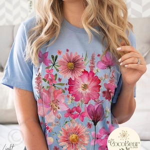May include: Pale blue t-shirt with a colourful floral print in pink and red hues. The design showcases a variety of flowers, giving a botanical feel. The words "Light Blue" are visible on the left. The CocoBear logo is on the right.