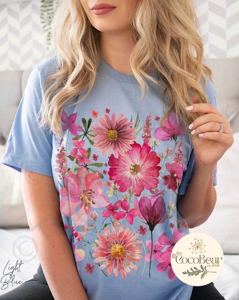 May include: Pale blue t-shirt with a colourful floral print in pink and red hues. The design showcases a variety of flowers, giving a botanical feel. The words "Light Blue" are visible on the left. The CocoBear logo is on the right.
