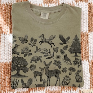 May include: Khaki-colored t-shirt with a nature-themed graphic. The design features detailed illustrations of woodland creatures, including a fox, deer, birds, and a rabbit, along with trees and flowers. The shirt has a classic crew neck.