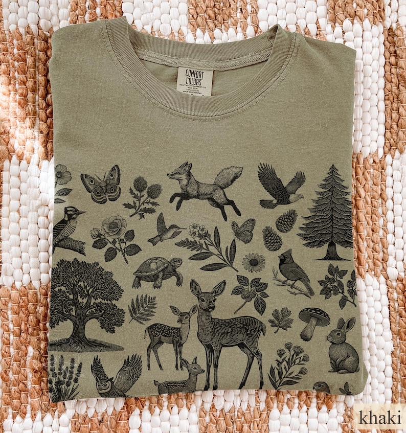 May include: Khaki-colored t-shirt with a nature-themed graphic. The design features detailed illustrations of woodland creatures, including a fox, deer, birds, and a rabbit, along with trees and flowers. The shirt has a classic crew neck.