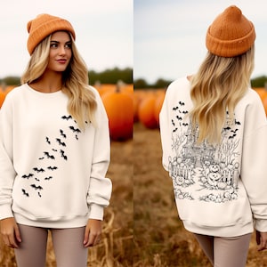 Op de afbeelding: Een wit sweatshirt met een zwart vleermuisontwerp aan de voorkant en een zwart spookhuisontwerp aan de achterkant. Het model draagt ​​een bruine beanie.