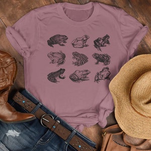 Puede incluir: Camiseta de color malva con una cuadr&iacute;cula de nueve ilustraciones detalladas de ranas en tinta negra. La camiseta se combina con un cintur&oacute;n de cuero marr&oacute;n, vaqueros, un sombrero de paja y botas de cuero marr&oacute;n.