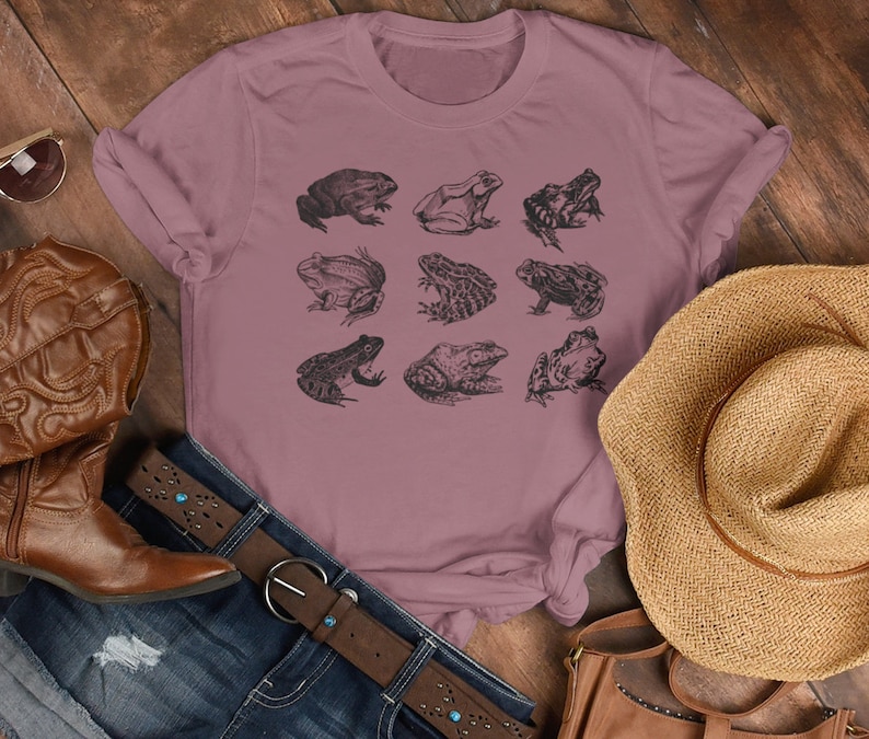 Puede incluir: Camiseta de color malva con una cuadr&iacute;cula de nueve ilustraciones detalladas de ranas en tinta negra. La camiseta se combina con un cintur&oacute;n de cuero marr&oacute;n, vaqueros, un sombrero de paja y botas de cuero marr&oacute;n.