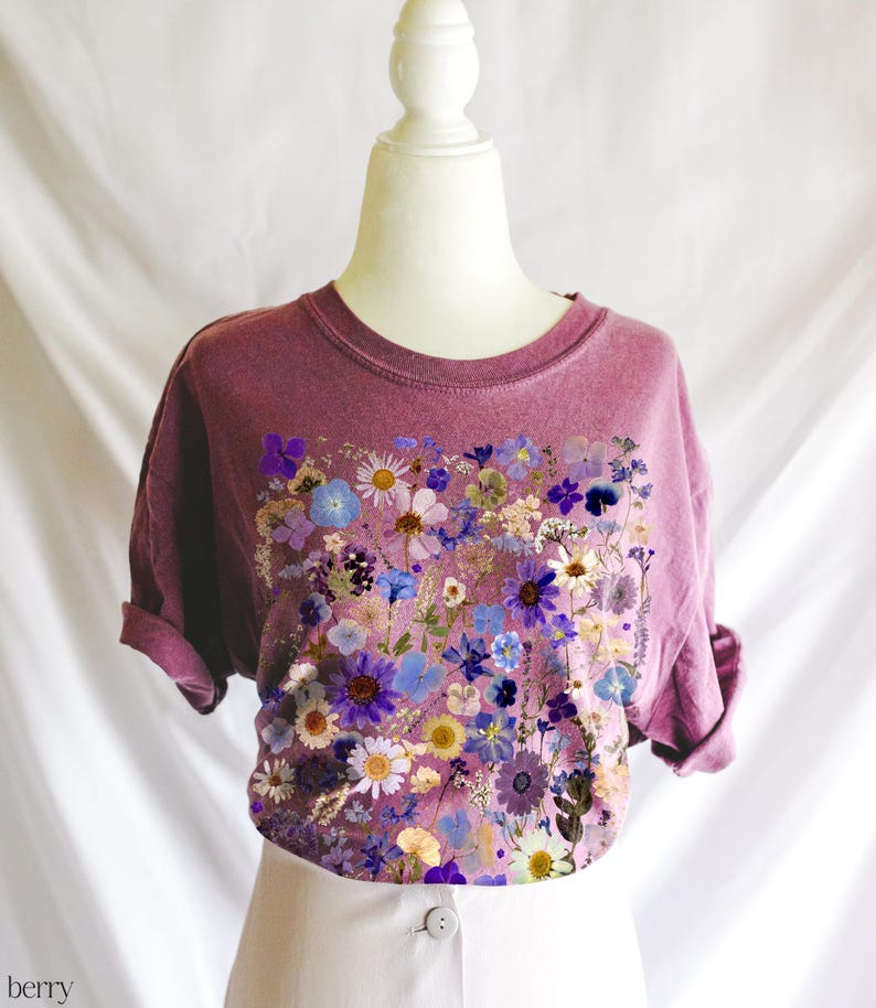 May include: A mauve t-shirt with a floral print design. The print features a variety of dried flowers in shades of purple, blue, and white.