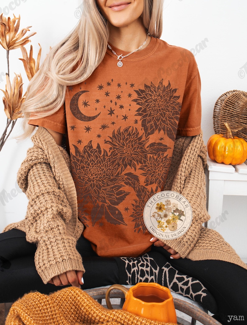Pode incluir: Uma camiseta cor de ferrugem com um design floral e celestial. O gr&aacute;fico inclui uma lua crescente, estrelas e grandes ilustra&ccedil;&otilde;es de flores em um tom marrom mais escuro. A camiseta faz parte da cole&ccedil;&atilde;o "The Coco Bear Original Design".