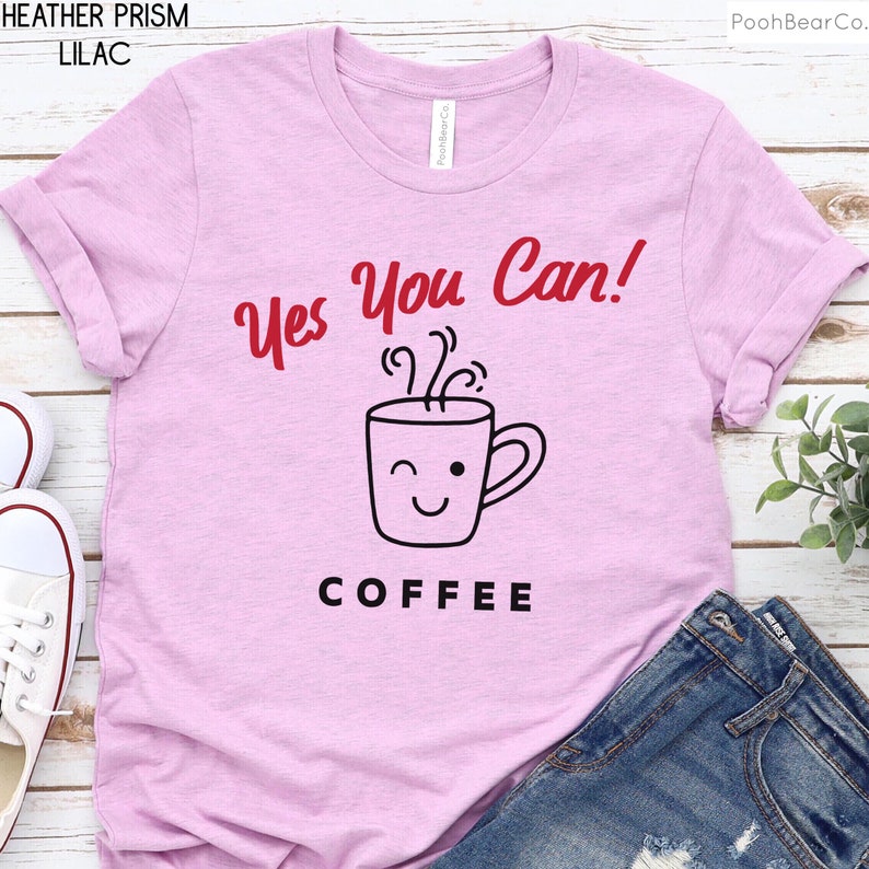 Może przedstawiać: Jasnofioletowa koszulka z czerwonym napisem "Yes You Can!" nad grafiką mrugającej filiżanki kawy. Poniżej filiżanki słowo "COFFEE" jest wydrukowane na czarno. Koszulka jest zestawiona z niebieskimi dżinsami.