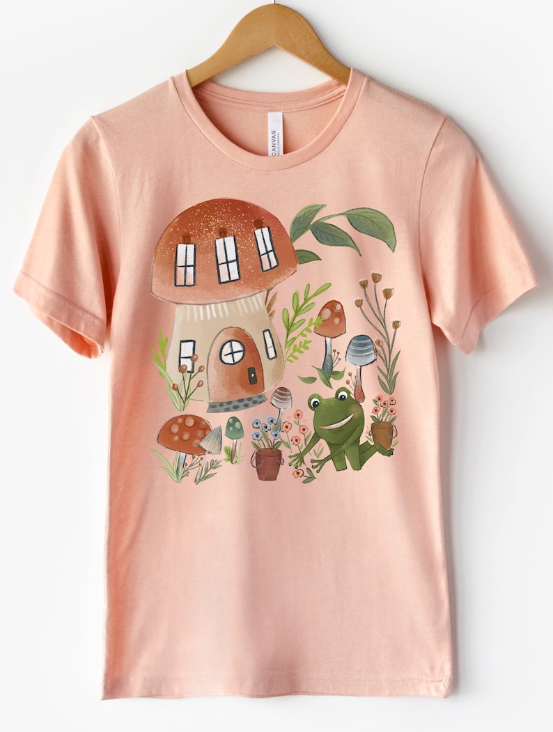 K&ouml;nnte beinhalten: Ein pfirsichfarbenes T-Shirt mit einer fantasievollen Illustration eines Pilzhauses, eines Frosches und Pilzen. Die Illustration ist im Cartoon-Stil mit leuchtenden Farben und Details gehalten.