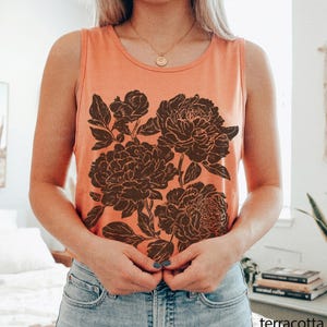 May include: A sleeveless tank top in a solid terracotta orange color featuring a large, dark brown graphic print of blooming peony flowers and leaves centered on the front. The garment has a round neckline and a relaxed, casual fit.