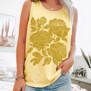 May include: A butter yellow sleeveless tank top featuring a large, detailed mustard yellow floral graphic print on the front. The design showcases multiple blooming flowers with intricate petals and leaves, centered on the chest of the garment.