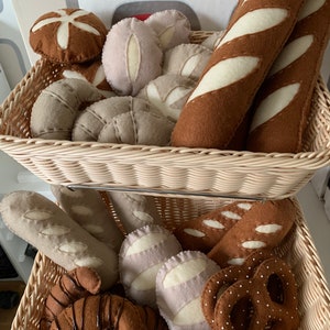 Könnte beinhalten: Ein Weidenkorb gefüllt mit Filz-Lebensmitteln, darunter ein Baguette, Croissants, Brezeln und verschiedene Brote. Der Korb ist auf einem weiteren Weidenkorb gestapelt, der mit weiteren Filz-Lebensmitteln gefüllt ist.