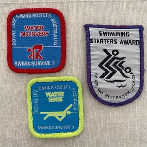 May include: Three swimming achievement badges. The badges feature text including "Water Discovery", "Swim & Survive 1", "Water Sense", and "Swim & Survive 3". The badges are in blue, red, purple, and green.