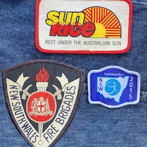 May include: A denim jacket with three patches sewn on. The top patch is white with red and yellow accents and says "Sun Rice" and "Best Under the Australian Sun". The left patch is black with white and red accents and says "New South Wales Fire Brigades". The right patch is blue with white accents and says "Gymnastics NSW 2015".